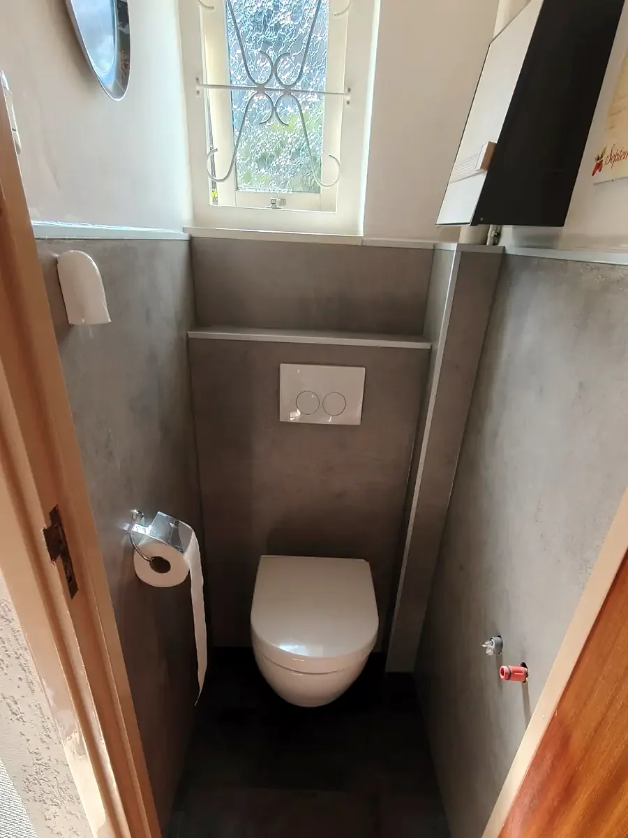 Hetzelfde smalle toilet na de renovatie: hangtoilet en grijze wandpanelen onder het sierraam